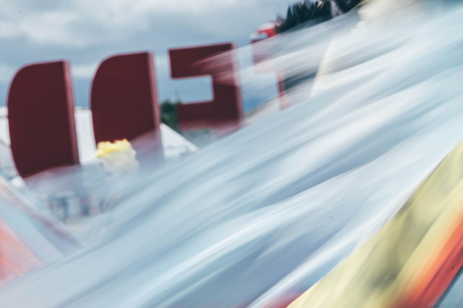 Blurred motion of colorful fabric or banner in the foreground with partial red letters in the background, likely part of an outdoor festival sign under a cloudy sky