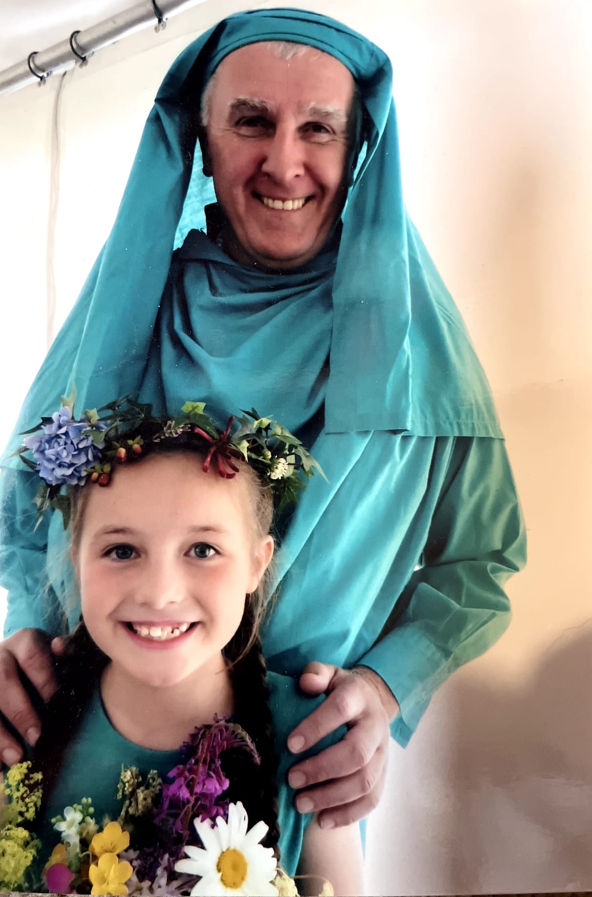 Two people standing together; one in the background wearing a green Gorsedd robe and head-dress, with hands resting on the shoulders of the person in front, who is wearing a teal dress and a colourful flower crown
