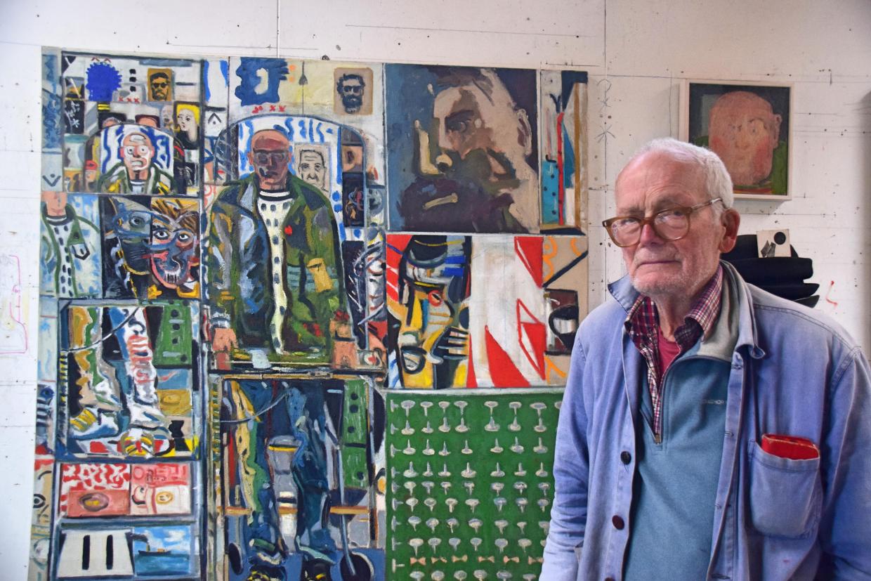 Man in glasses and a blue jacket with a red pocket square standing in front of a large colorful painting with abstract and figurative elements; a smaller portrait hangs on the wall to the right