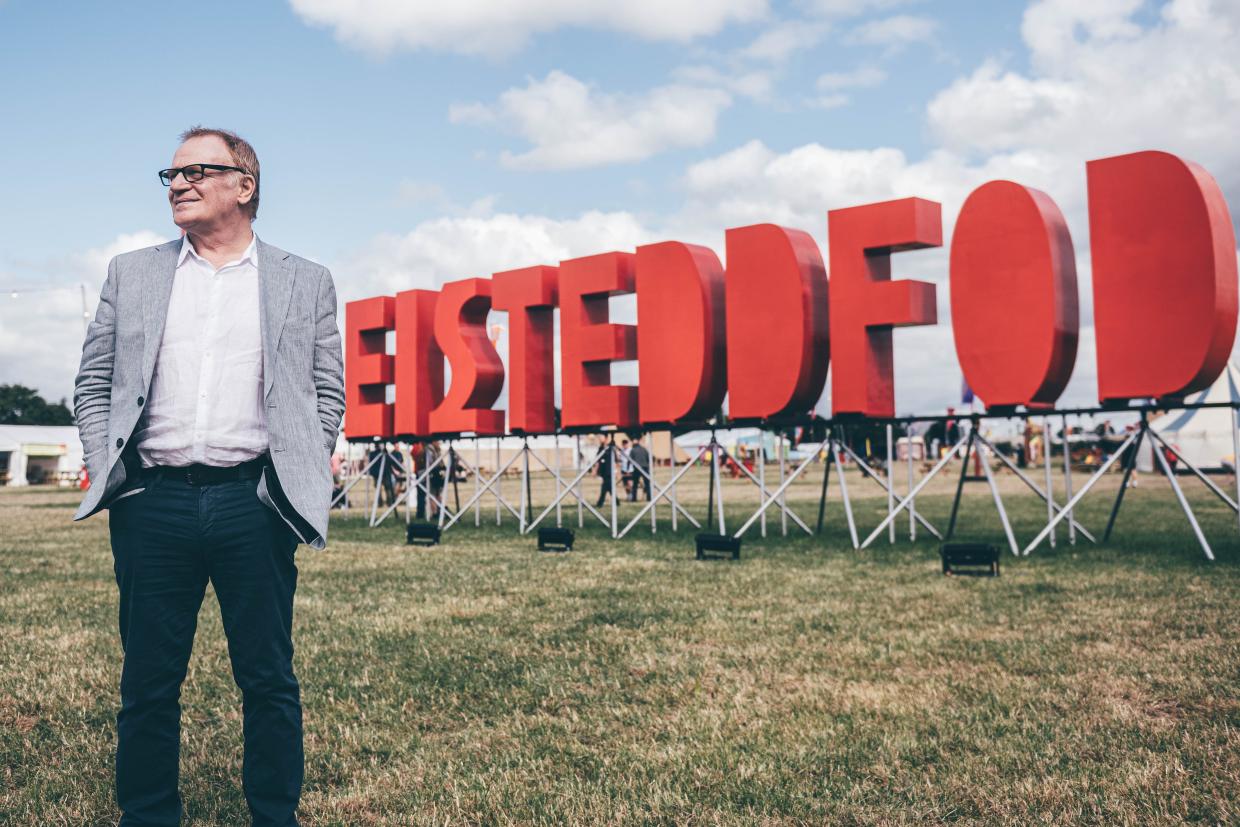 Man in a light gray blazer and dark pants standing on grass in front of a large red 'EISTEDDFOD' sign, with people and tents in the background under a partly cloudy sky
