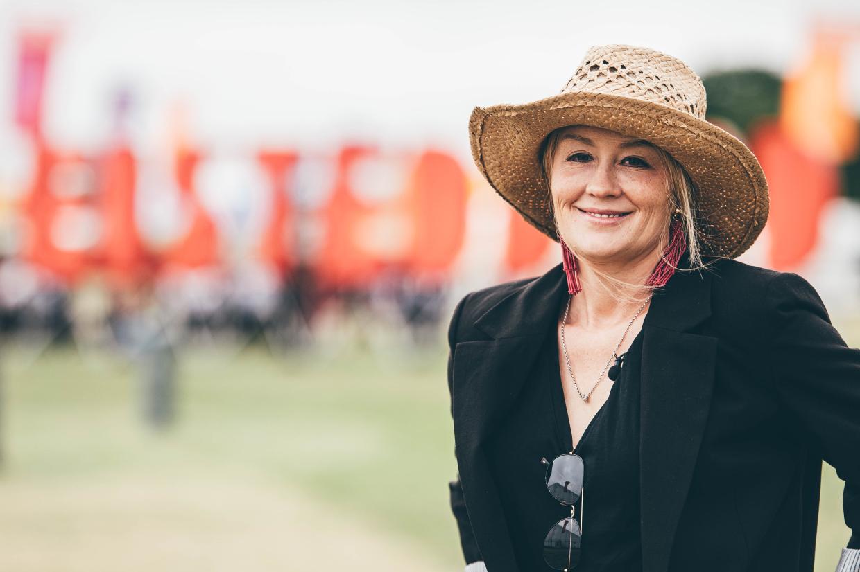 Person wearing a straw hat, black blazer, and sunglasses hanging from the neckline, standing outdoors with red banners and people in the blurred background