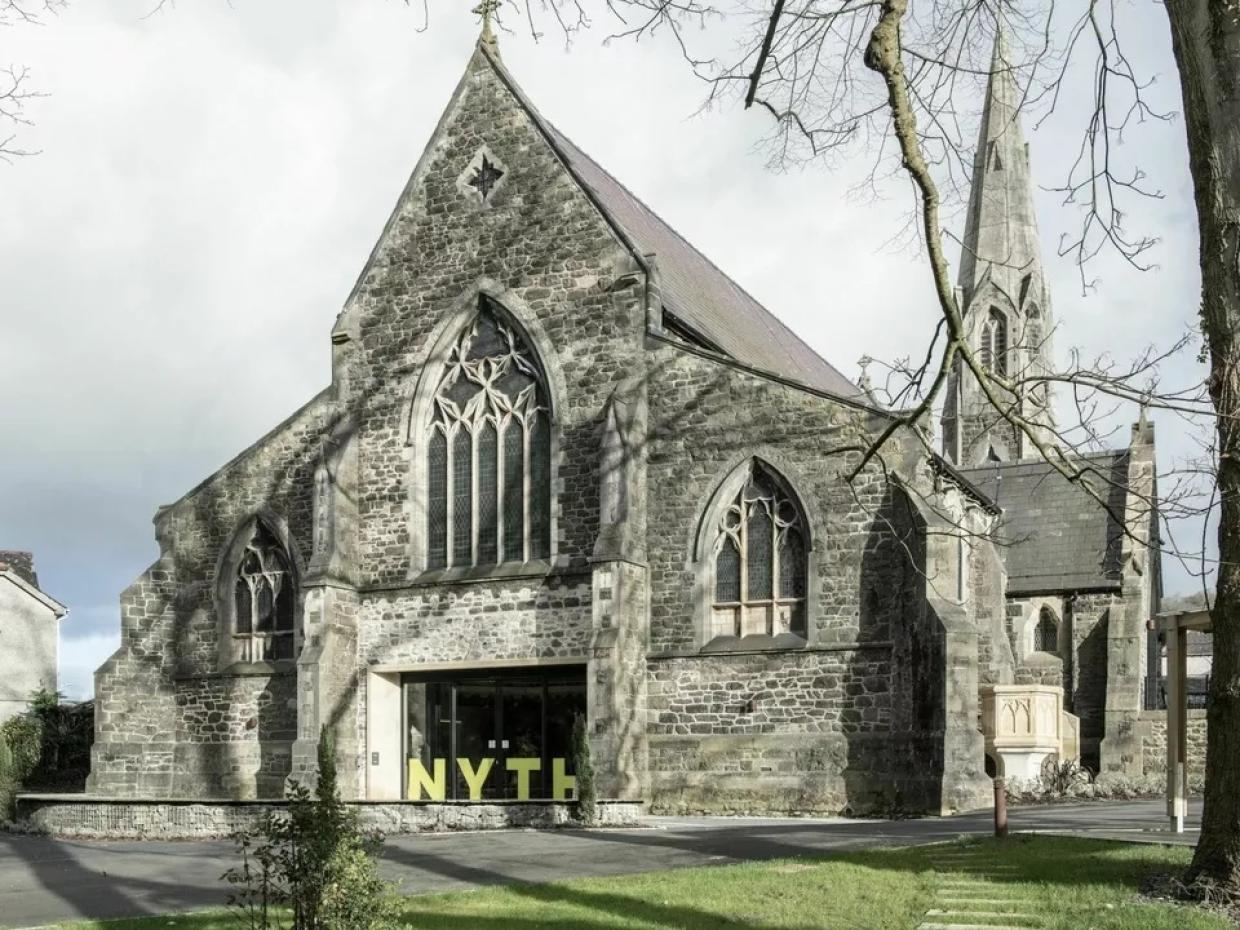 Exterior of a stone Gothic-style church with pointed arches and large stained glass windows; modern glass entrance doors display the word 'NYTH' above them, with a tall spire and greenery in the background