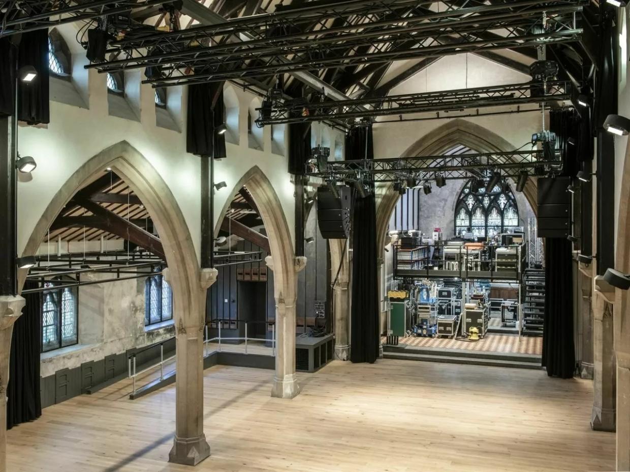 Interior of a large gothic-style hall with pointed arches, high ceilings, and light wooden flooring; a stage with black curtains and technical equipment is set up at one end, with lighting rigs and speakers suspended above
