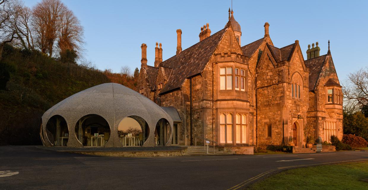 Historic stone building with steep roofs and multiple chimneys, next to a modern dome-shaped extension with large arched windows; sunset light casts a warm glow over the scene, with trees in the background and a paved driveway in front