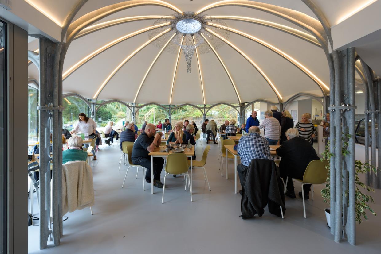 Indoor dining area with a dome-shaped ceiling and central chandelier; people seated at green and white chairs around tables, eating and socialising; large windows in the background reveal greenery outside