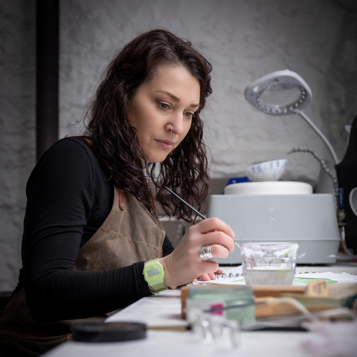Person wearing a brown apron and black shirt working at a desk with craft tools and materials; face blurred for privacy. A magnifying lamp and glass of water are also visible