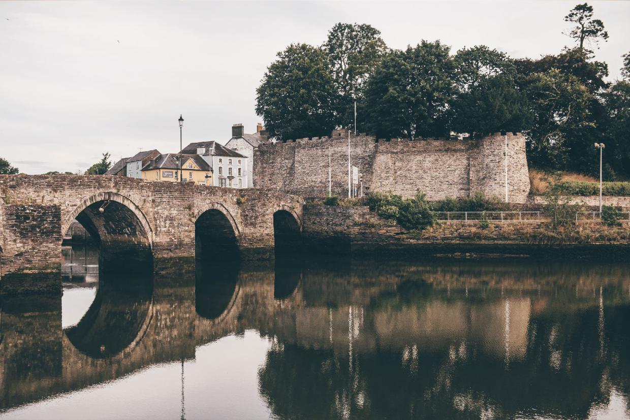 Old stone bridge with three arches reflected in calm water, with trees, stone wall, and buildings in the background