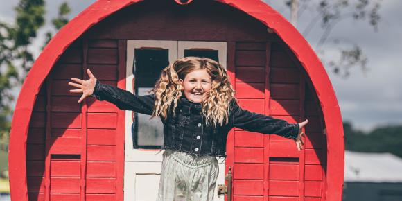 Young girl with long blonde hair hanging out of a red wooden caravan in a field with a cloudy sky