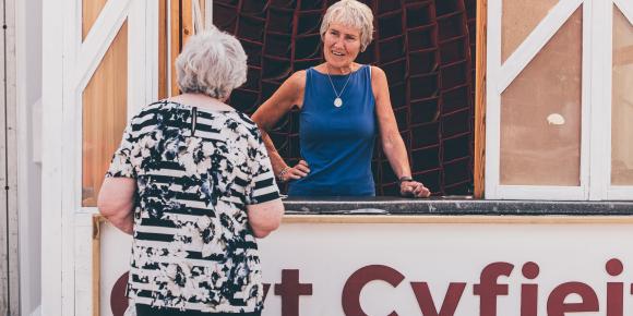 Two people interacting at a wooden booth labeled 'Cwt Cyfieithu Translation Hut'; one person stands inside behind a glass window, and the other stands outside facing the booth