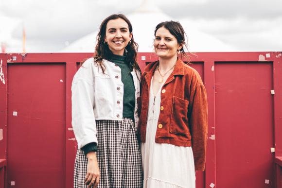 Two individuals standing in front of a red wall under a cloudy sky; one wearing a white jacket and checkered skirt, the other in an orange-brown jacket and white dress