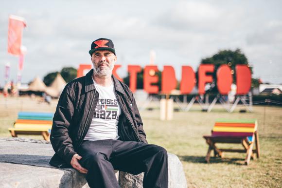 Person wearing 'Artists for Gaza' t-shirt seated on a stone bench outdoors, with large red 'EISTEDDFOD' letters and colourful benches and tents in the background