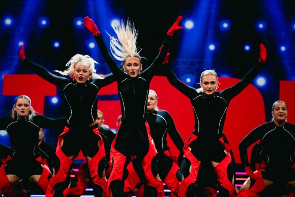 Group of performers in black and red costumes with red gloves dancing energetically on stage under bright lights, with large red letters in the background