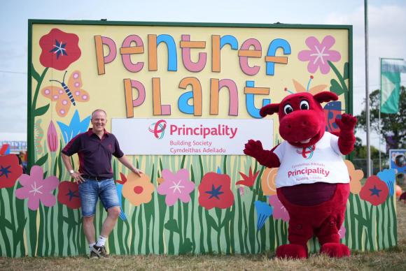 Colorful 'Pentref Plant' sign with flowers and a butterfly, featuring the Principality Building Society logo; a person stands on the left and a red dragon mascot wearing a 'Principality' shirt on the right