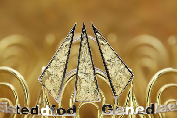 Close-up of a golden metallic structure with three central triangular shapes, blurred golden background, and text 'Eisteddfod Genedlaethol' at the bottom