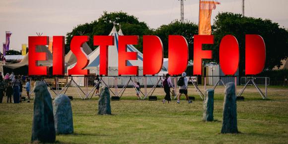 Digwyddiad awyr agored gyda llythrennau 'EISTEDDFOD' mawr o liw coch, pobl yn cerdded ymhlith gwersylloedd a phlanhigion, a chylch yr Orsedd.