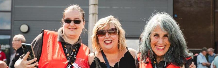 Tair merch mewn siacedi llachar oren yn gwirfoddoli gyda'r Eisteddfod