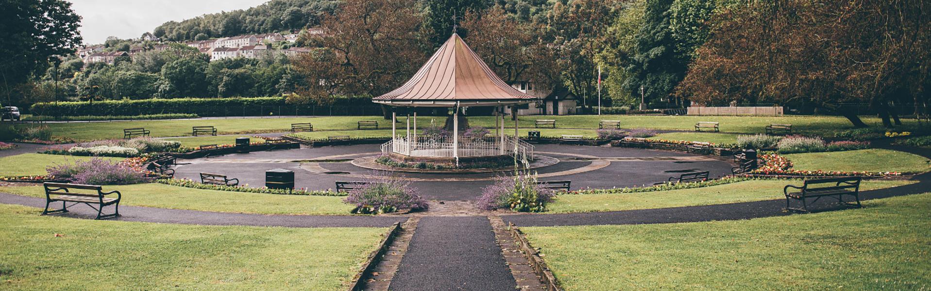 Bandstand yng nghanol Parc Ynysangharad, Pontypridd, wedi'i amgylchynnu ganu welyau blodau tymhorol ac ardal suddedig ffufiol ar gyfer cynulleidfa