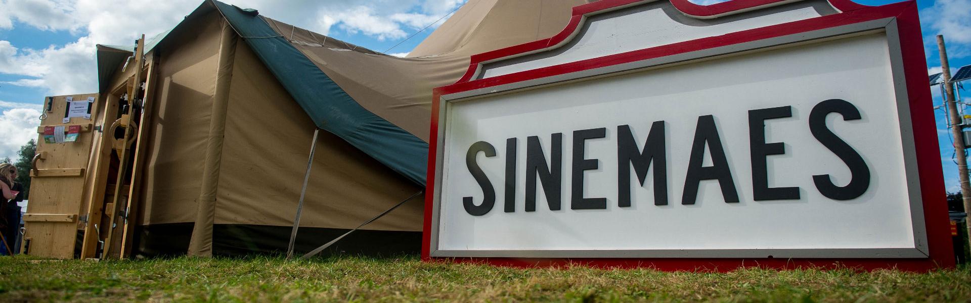 Arwydd Sinemaes mewn sgwar coch wrth ymyl tepee ar Faes yr Eisteddfod mewn tywydd braf