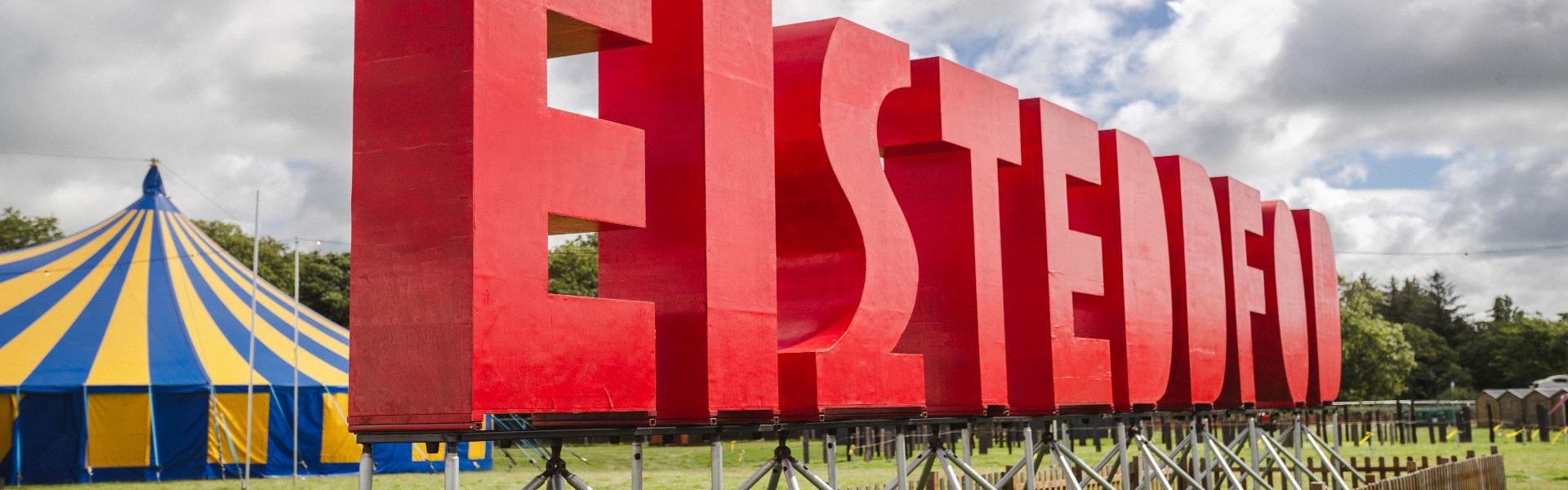 Llythrennau coch enfawr yn sillafu 'Eisteddfod' ar ongl mewn cae gyda phafiliwn streipiog yn y cefndir ac awyr gymylog uwchben