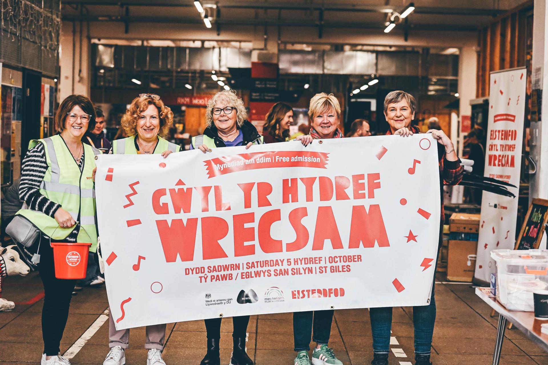 5 o ferched y tu ôl i faner wen gyda geiriau Gwyl yr Hydref Wrecsam'' yn gwisgo siacedi llachar ar gefndir prysur mewn adeilad