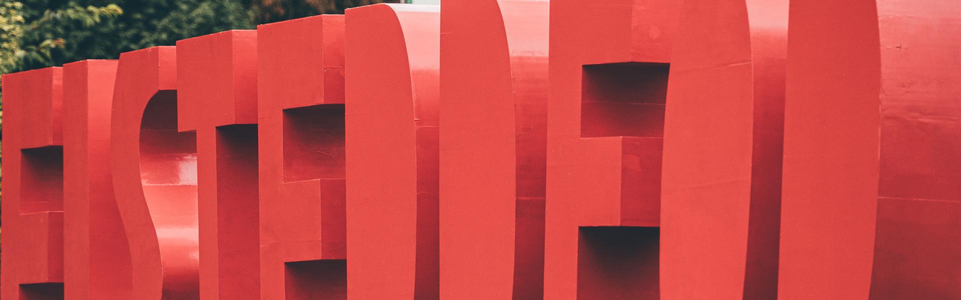 Large red 3D letters spelling 'EISTEDDFOD' displayed outdoors on a platform, with trees and a green and white flag in the background