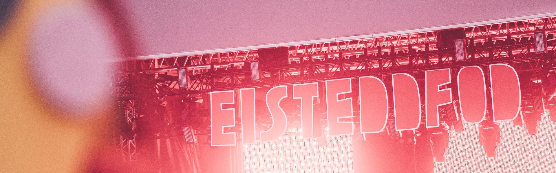 Outdoor stage with large 'EISTEDDFOD' sign, illuminated by red lights, and trees in the background. Blurred foreground object on the left