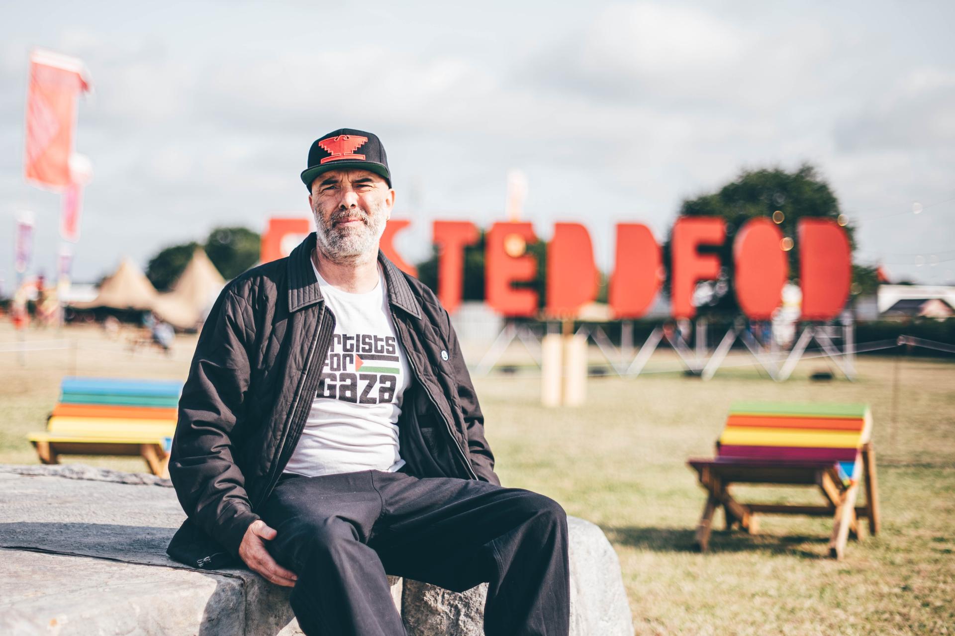 Person wearing 'Artists for Gaza' t-shirt seated on a stone bench outdoors, with large red 'EISTEDDFOD' letters and colourful benches and tents in the background