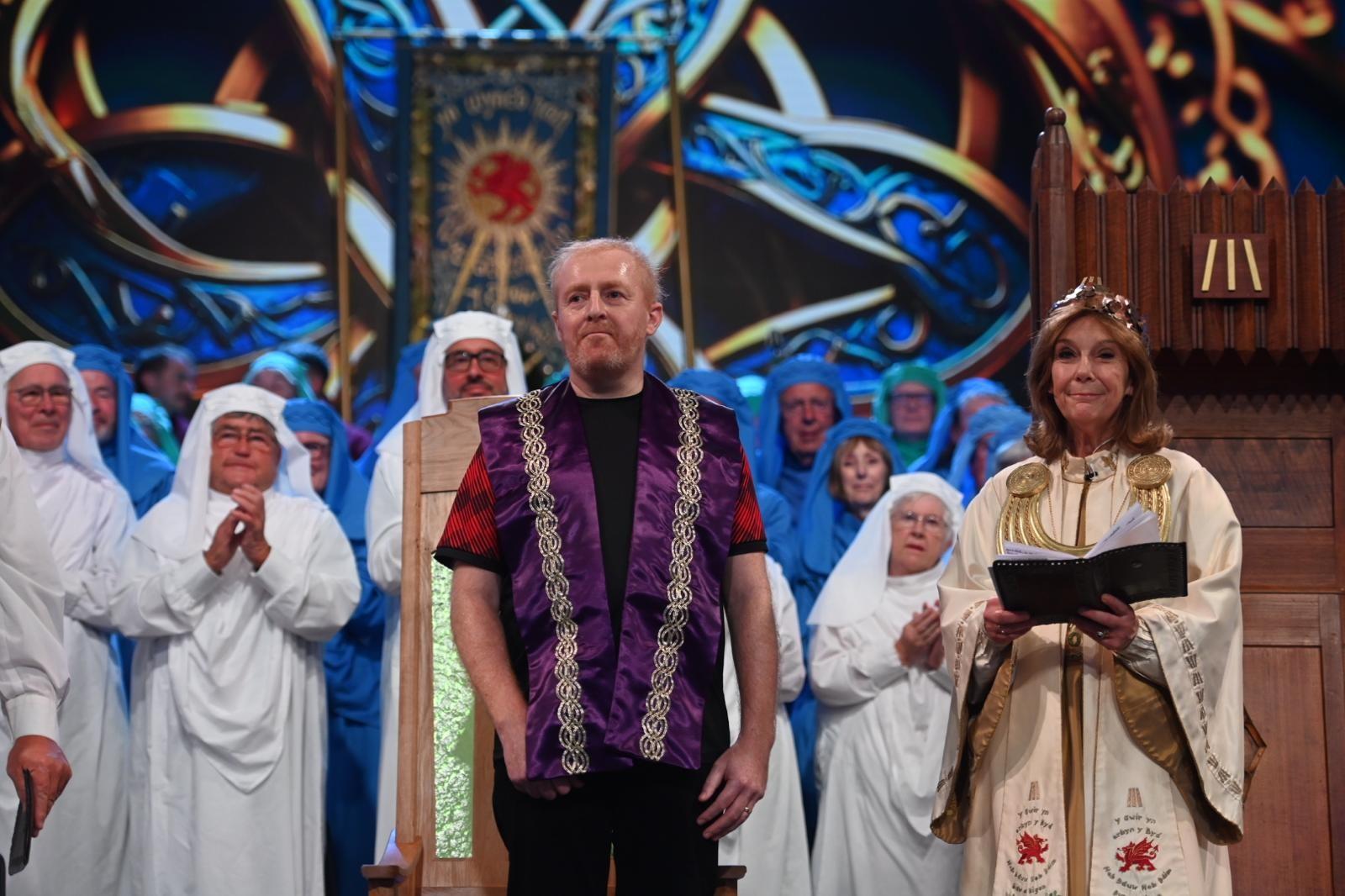 Group of people in ceremonial attire, including two central figures in ornate robes—one in purple and gold, the other in white with red symbols holding an open book—set against a backdrop with intricate designs and a sun emblem banner