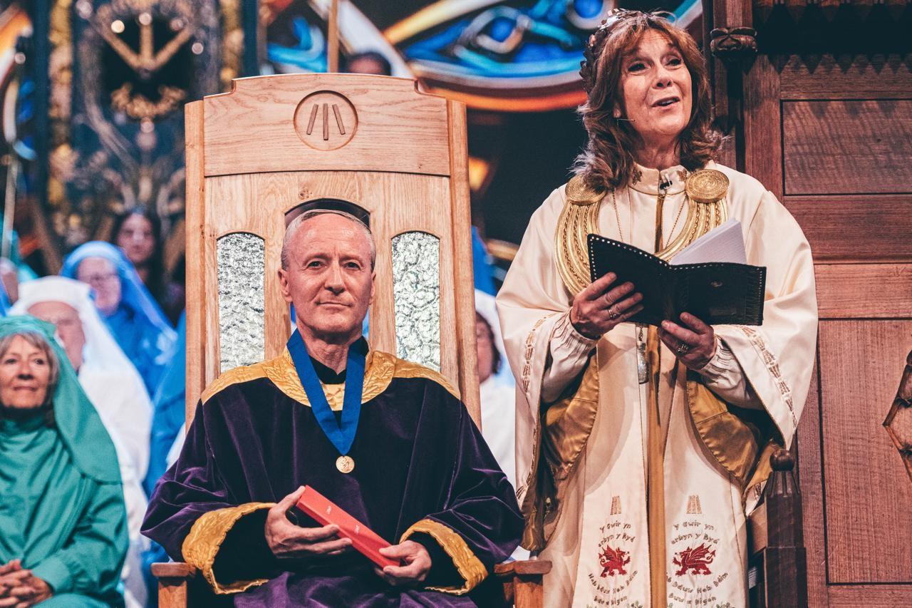 Ceremonial scene with a seated figure in a purple robe holding a red book, and a standing figure in white and gold attire holding an open folder, with others in the background at a formal event