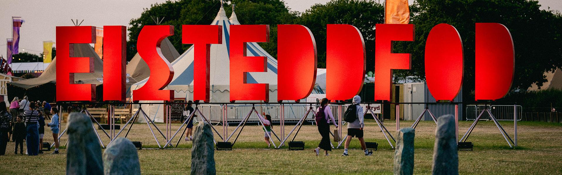 Digwyddiad awyr agored gyda llythrennau 'EISTEDDFOD' mawr o liw coch, pobl yn cerdded ymhlith gwersylloedd a phlanhigion, a chylch yr Orsedd.